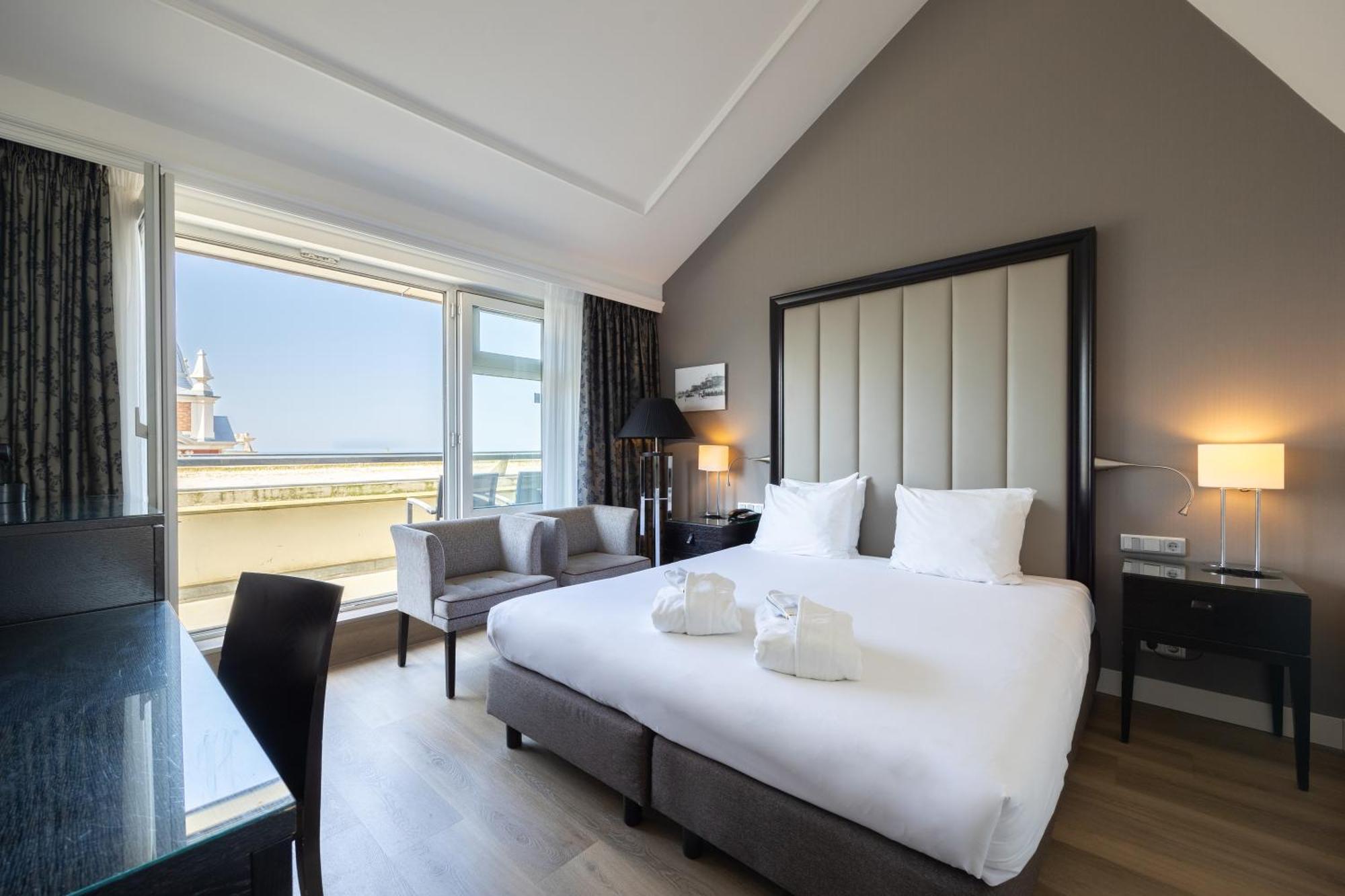 Grand Hotel Amrath Kurhaus The Hague Scheveningen Экстерьер фото The photo shows a modern hotel room featuring a spacious layout. There's a large bed with white linens and two decorative towels arranged on it. Next to the bed, there are two bedside lamps and a small table. A seating area is visible with two chairs